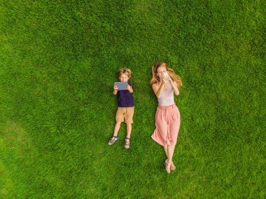 Anne ve oğlu parkta çimlerin üzerinde uzanıyorlar. Anne telefona bakar, oğul tablete bakar. Drone, quadracopter 'dan fotoğraflar