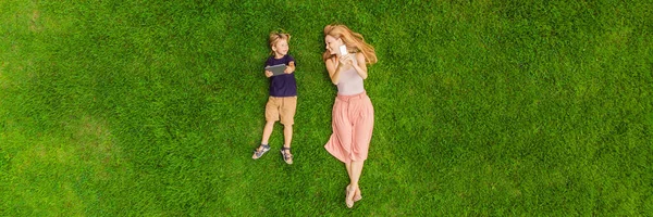 Mom and son are lying on the grass in the park. Mom looks at the phone, son looks at the tablet. Photos from the drone, quadracopter BANNER, LONG FORMAT