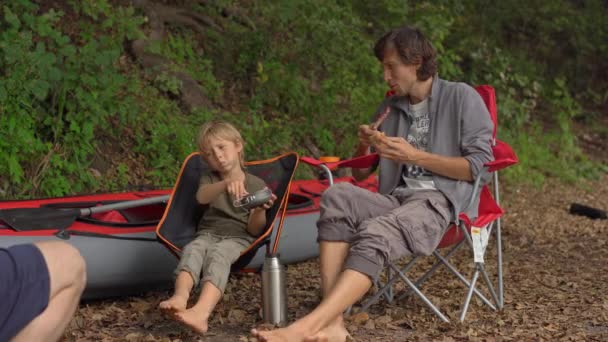 A young man and his little son have a break by a bonfire. They sit in front of a big inflatable kayak — ストック動画