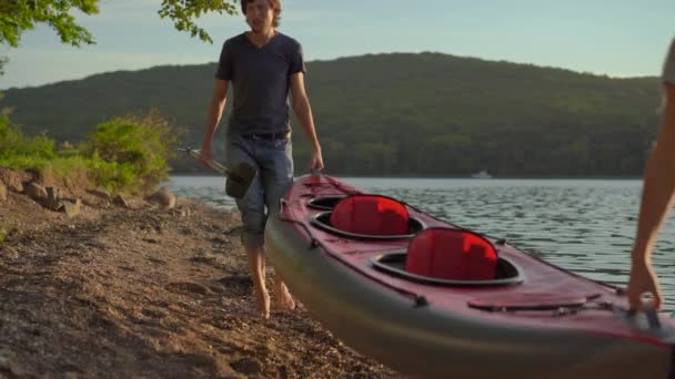 Un joven hombre y una mujer turistas llevando un gran kayak inflable junto a una orilla del mar o un lago — Vídeos de Stock