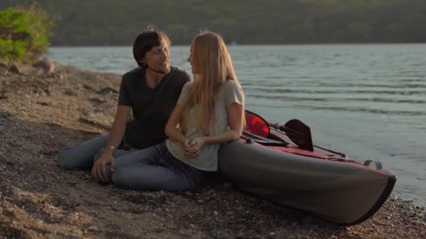 Un joven hombre y una mujer turistas sentados junto a un gran kayak inflable en una orilla del mar o un lago — Vídeo de stock
