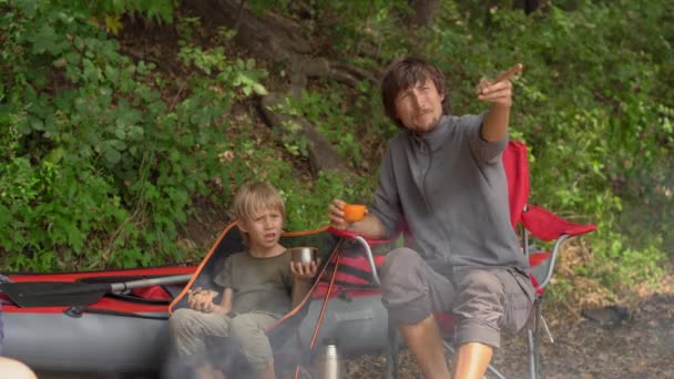 Un hombre joven y su hijo pequeño tienen un descanso junto a una hoguera. Se sientan frente a un gran kayak inflable — Vídeos de Stock