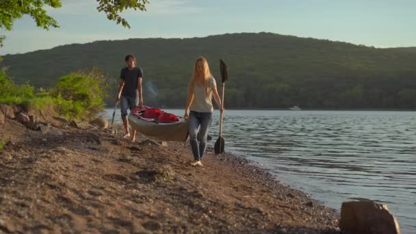 Egy fiatal férfi és egy nő turista, akik egy nagy felfújható kajakot cipelnek egy tengerparton vagy egy tónál. — Stock videók