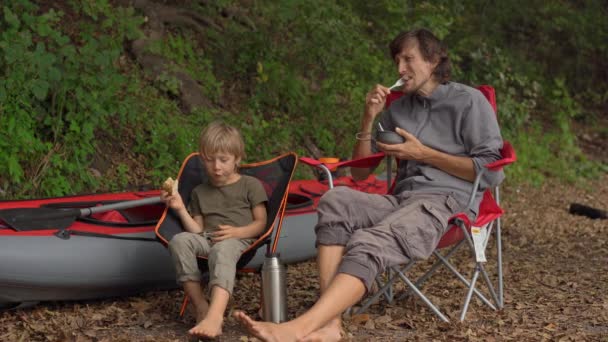 一个年轻人和他的小儿子在篝火边休息一下。 他们坐在一个充气的大皮划艇前 — 图库视频影像