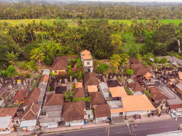 Many villas with brown-orange shingle roofs between tropical trees on the sky background in Ubud on Bali. Sun is shining onto them. Aerial horizontal photo