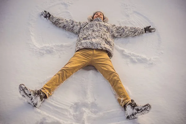 Молодий чоловік у куртці лежить на снігу і робить фігуру ангела. Зимові забави — стокове фото