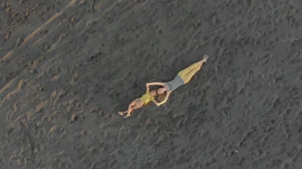 Tiro aéreo de uma mãe de família e seu filho deitado em uma praia de areia preta — Vídeo de Stock