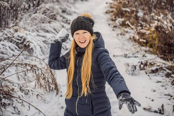 Зимняя девушка бросает снежок в камеру, улыбаясь счастливо веселясь на открытом воздухе в снежный зимний день, играя в снегу. Милая игривая молодая женщина на открытом воздухе наслаждается первым снегом — стоковое фото