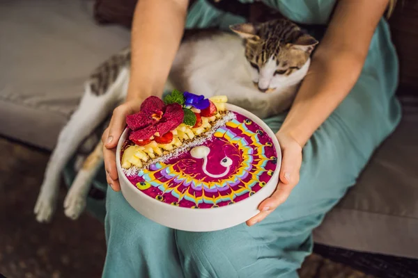 Молодая женщина, завтракающая в средиземноморском стиле на своем диване и со своим котом, ест здоровый тропический завтрак, чашу смузи с тропическими фруктами, украшенную рисунком красочного йогурта — стоковое фото