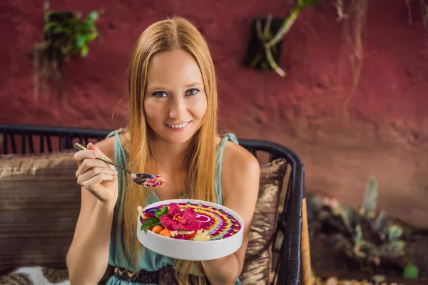 Young woman having a mediterranean breakfast seated at her sofa and eats Healthy tropical breakfast, smoothie bowl with tropical fruits, decorated with a pattern of colorful yogurt with turmeric and — Stock Photo, Image