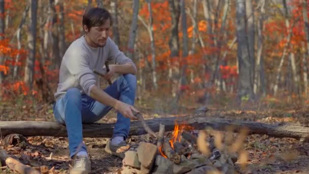 Young man sitting by a bonfire in a cold autumn forest. Autumn vacation concept — Stock Video