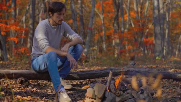 Un joven sentado junto a una hoguera en un frío bosque otoñal. Concepto de vacaciones de otoño — Vídeo de stock