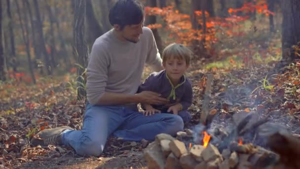 El joven y su pequeño hijo sentados junto a una hoguera en un frío bosque otoñal. Concepto de vacaciones de otoño — Vídeo de stock