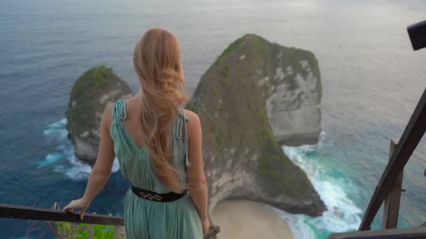若い女性の観光客は、インドネシアのヌサペニダ州ケリングキングビーチにあるいわゆるティラノサウルス岩を見ることができます。ヌサペニダ島の有名な観光地 — ストック動画