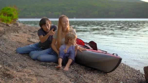 Une jeune famille - le père, la mère et le fils s'assoient à côté de leur kayak gonflable sur un lac ou un rivage. Coup de ralenti — Video