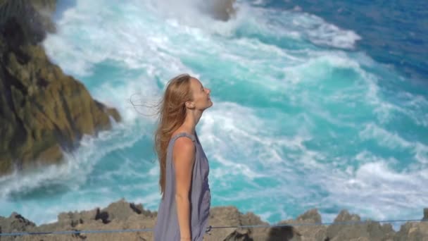 若い女性の観光客は、巨大な波が岩に打ち寄せているヌサペニダ島のエンジェルスビラボンビーチを訪れます。スローモーションショット — ストック動画