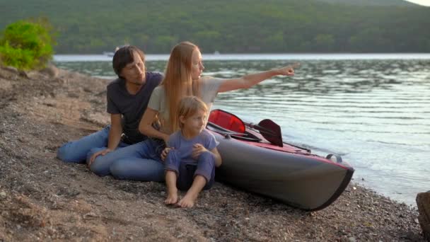 Молодая семья - отец, мать и сын сидят у надувного каяка на озере или на берегу моря. Слоумоушн — стоковое видео