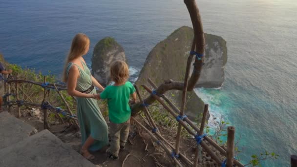 La joven y su pequeño hijo turistas visitan la llamada roca Tiranosaurio en la playa Kelingking, Nusa Penida, Indonesia. Famoso lugar turístico en la isla de Nusa Penida. Disparo en cámara lenta — Vídeos de Stock