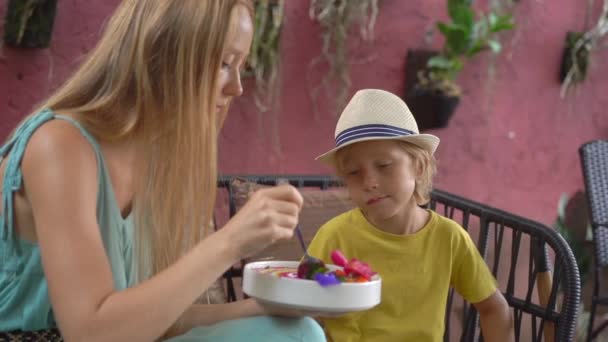 Giovane donna e il suo piccolo figlio godono colorato frullato di muesli nella ciotola seduto in un bellissimo caffè. Un concetto di alimentazione sana. Colpo di rallentamento — Video Stock