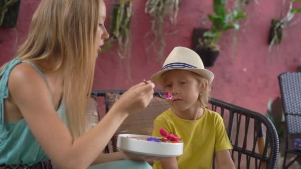 Mujer joven y su pequeño hijo disfrutan de colorido batido de granola en el tazón sentado en un hermoso café. Concepto de alimentación saludable. Disparo en cámara lenta — Vídeo de stock