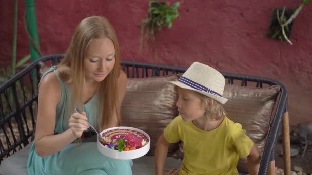 Young woman and her little son enjoy colorful granola smoothie in the bowl sitting in a beautiful cafe. Healthy eating concept. Slowmotion shot — ストック動画