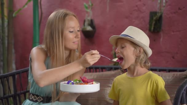 Mujer joven y su pequeño hijo disfrutan de colorido batido de granola en el tazón sentado en un hermoso café. Concepto de alimentación saludable. Disparo en cámara lenta — Vídeos de Stock