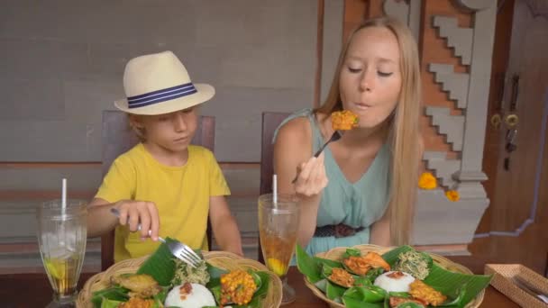 Young woman and her little son eat traditional Balenese food set Nasi Campur in a traditional cafe. Travel to Bali concept — ストック動画