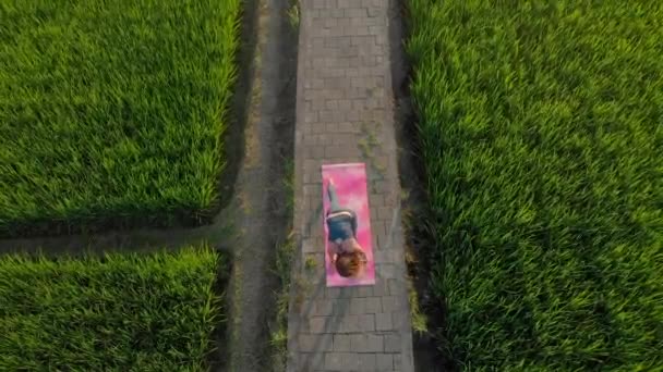 Повітряний постріл молодої жінки, яка під час заходу сонця практикує йогу на великому полі з прекрасним рисом. Подорож до Азії. — стокове відео