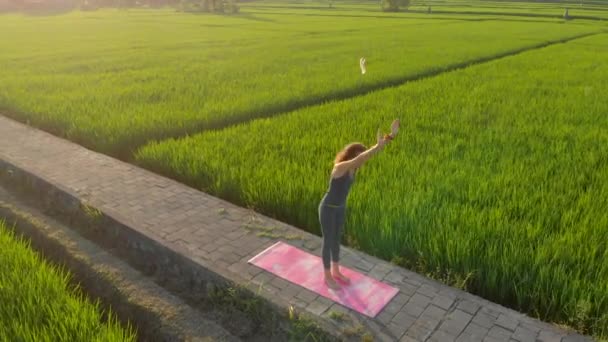 Εναέρια λήψη μιας νεαρής γυναίκας που κάνει γιόγκα σε ένα μεγάλο όμορφο χωράφι ρυζιού κατά τη διάρκεια του ηλιοβασιλέματος. Ταξίδι στην Ασία έννοια. — Αρχείο Βίντεο