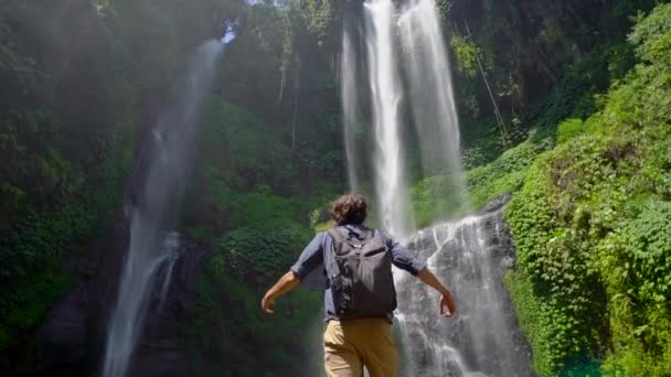 Young man tourist visits the biggest waterfall on the Bali island - the Sekumpul waterfall. Slowmotion shot. Travel to Bali concept — Stock Video