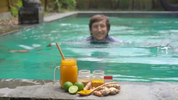 一个年轻人喝着民族巴厘人的酒，Jamu在游泳池里游泳 — 图库视频影像