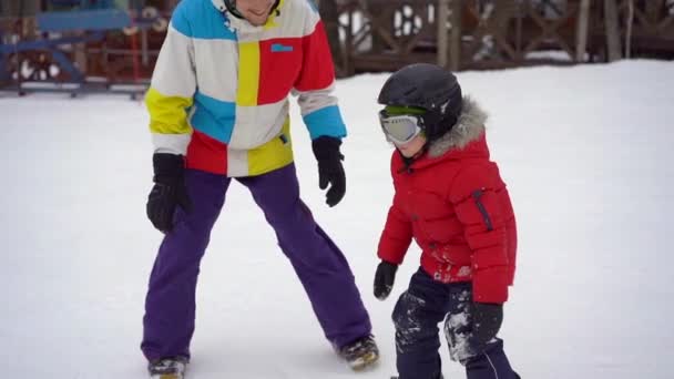 若い男のスノーボードインストラクターは小さな男の子にスノーボードに乗る方法を教えます。冬の活動の概念。スローモーションショット — ストック動画