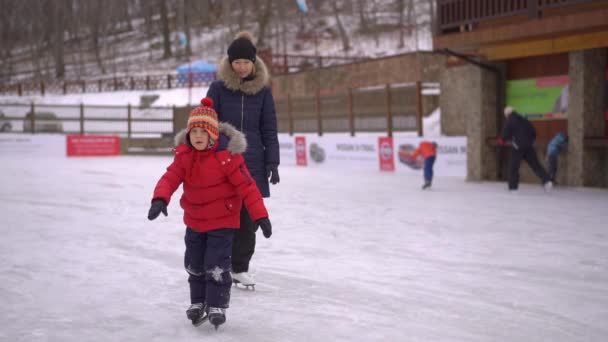 Una madre joven y un niño pequeño en una pista. El niño aprende a patinar sobre hielo. Concepto de actividades de invierno — Vídeos de Stock