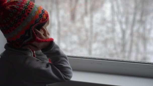 Ein kleiner Junge blickt durch das Fenster auf einen heftigen Schneefall, der auf Weihnachten wartet — Stockvideo