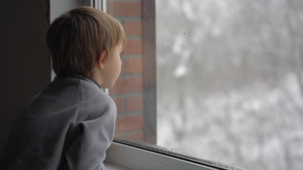 Un niño pequeño mira por la ventana en una fuerte nevada esperando que llegue la Navidad — Vídeos de Stock