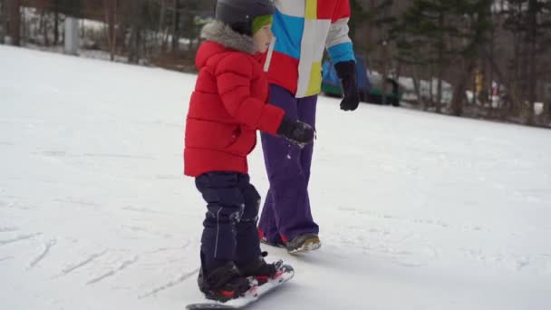 Молодой человек инструктор по сноуборду дает советы маленькому мальчику, как ездить на сноуборде. Концепция зимних мероприятий — стоковое видео