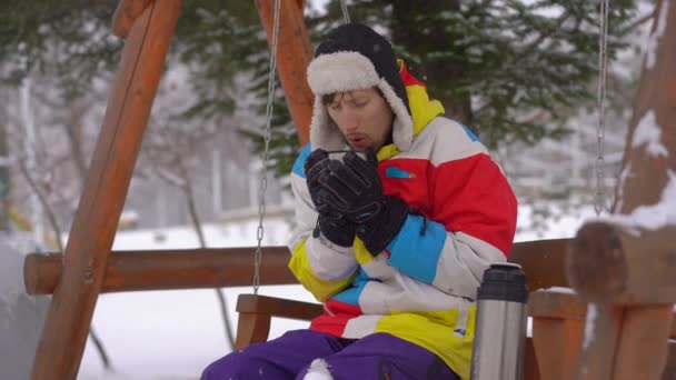 O jovem durante a queda de neve senta-se em um banco e bebe bebida quente. Ele sente muito frio. Conceito de tempo frio — Vídeo de Stock