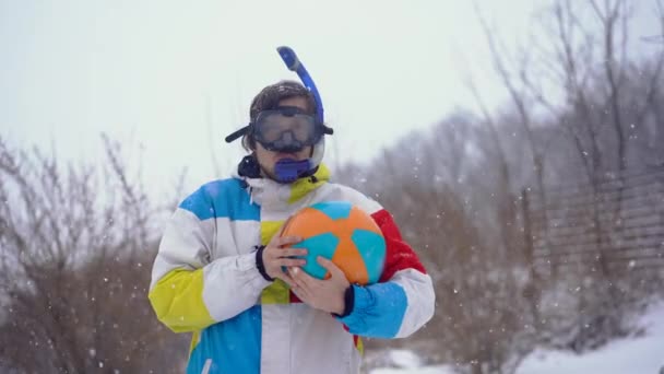 Забавный человек в водолазной маске с надувным мячом стоит под снегопадом. Опоздали во времени — стоковое видео