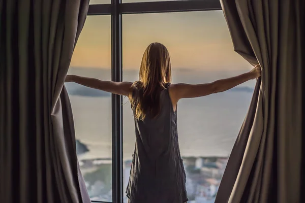 Jonge vrouw opent de gordijnen op een venster met uitzicht op zee — Stockfoto
