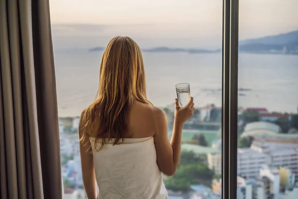 Donna beve acqua al mattino su uno sfondo di una finestra con vista sul mare — Foto Stock