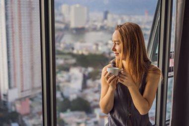 Genç bir kadın sabah kahve içiyor. Deniz manzaralı bir pencerenin arkasında.