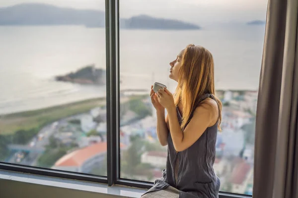 Giovane donna sta bevendo caffè al mattino sullo sfondo di una finestra con vista sul mare — Foto Stock