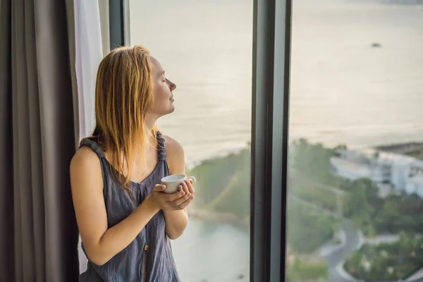 Молода жінка п'є каву вранці на фоні вікна з видом на море — стокове фото