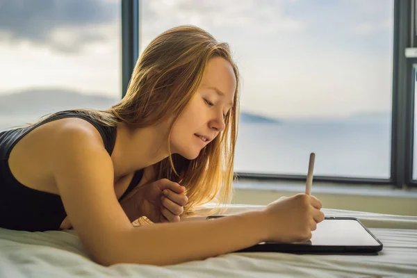 A fiatal nő rajzol egy tablettát a háttérben egy ablak kilátással a tengerre. Tervező, rajzolás — Stock Fotó