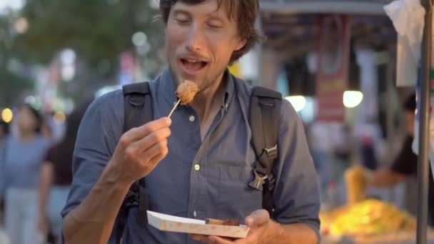 Junger Mann besucht den traditionellen koreanischen Straßenmarkt. Er isst Fischkuchen auf einem Bambusstab. Reise nach Korea. — Stockvideo