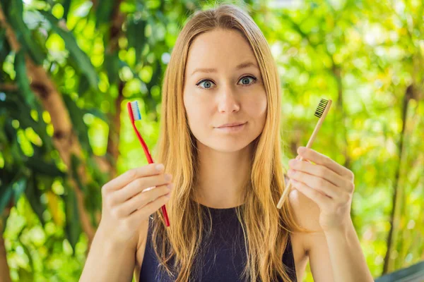 Donna tiene eco spazzolino da denti in bambù naturale e in altra mano spazzolino da denti in plastica. sfondo rustico. concetto di stile di vita sostenibile. zero sprechi. articoli liberi di plastica scelta — Foto Stock