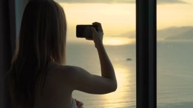 Sahilde ve gündoğumunda panoramik manzaralı bir dairede genç bir kadın. Güzel bir gün doğumunun fotoğrafını çeker.