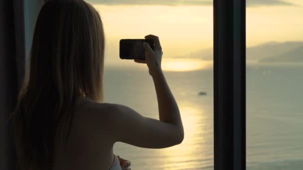 Een jonge vrouw in een appartement met een panoramisch uitzicht op de kust en zonsopgang. Ze maakt een foto van een mooie zonsopgang — Stockvideo