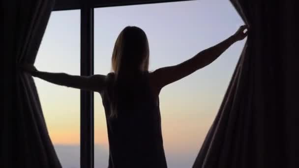 Uma jovem mulher em um apartamento com vista panorâmica sobre a costa e nascer do sol. Ela abre cortinas de janela — Vídeo de Stock