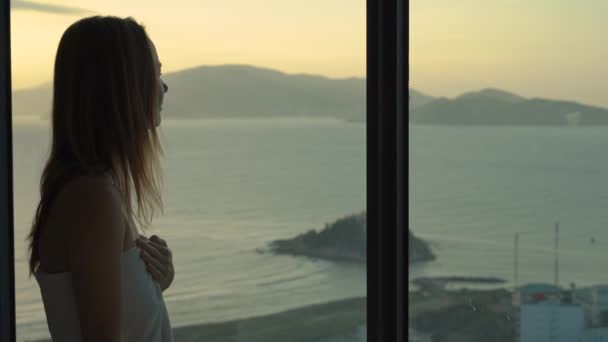 A young woman in an apartment with a panoramic view on the coast and sunrise. She uses her smarphone standing by the window — Stock Video